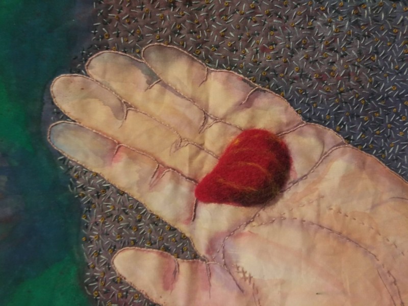 A red felted rock sits on an open palm filling the composition. The fabric is painted with a background of purple and green and embroidered with seed stitch and an outline around the hand