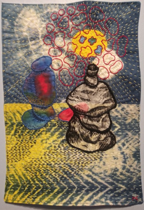 two rock cairns balance side by side, one black and white and on in color on a yellow, white, and blue background of tie-dyed patterns. A yellow circle floats behind them covered by red stitched rings. The shapes are outlined in satin stitch and the foreground is covered by rows of running stitch.